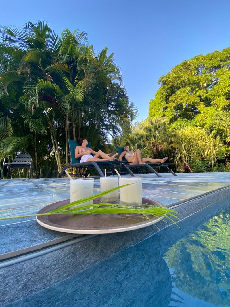 Deux personnes sur des chaises longues au bord de la piscine extérieur avec un plateau de boissons du bar du Caraïb'Bay Hôtel
